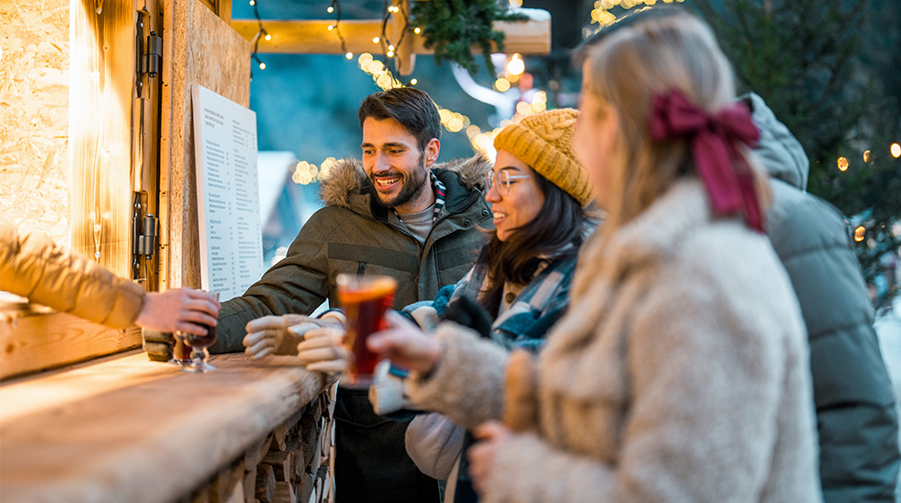 Candid portrait of diverse young couples socializing at wooden market counter, sharing mulled wine beneath twinkling string lights.
